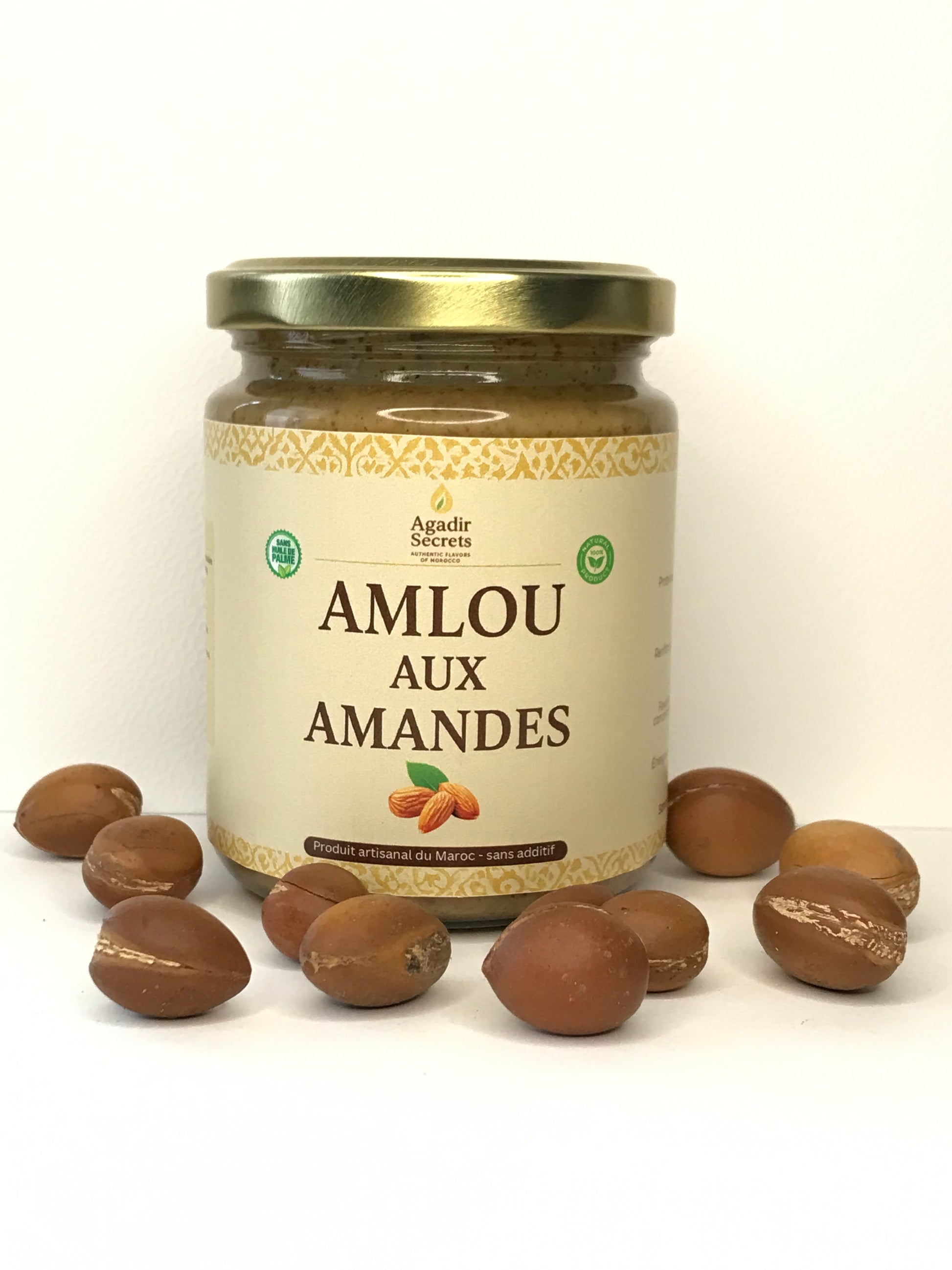 Pot en verre d'Amlou aux amandes Agadir Secrets entouré de véritables noix d'argan sur fond blanc. Une pâte à tartiner marocaine 100% naturelle et sans additifs, disponible en expédition rapide dans toute la France.